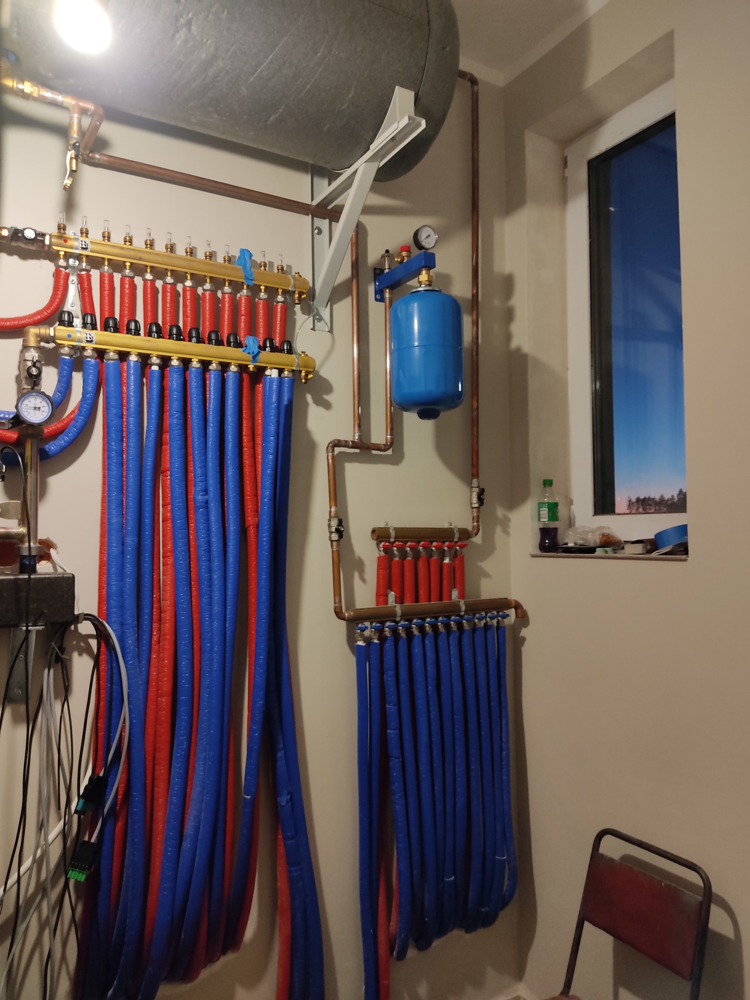 Rozdzielacz ogrzewania podłogowego z rurami w izolacji czerwonej i niebieskiej, zamontowany na ścianie w pomieszczeniu z oknem, widoczny manometr i zbiornik wyrównawczy.