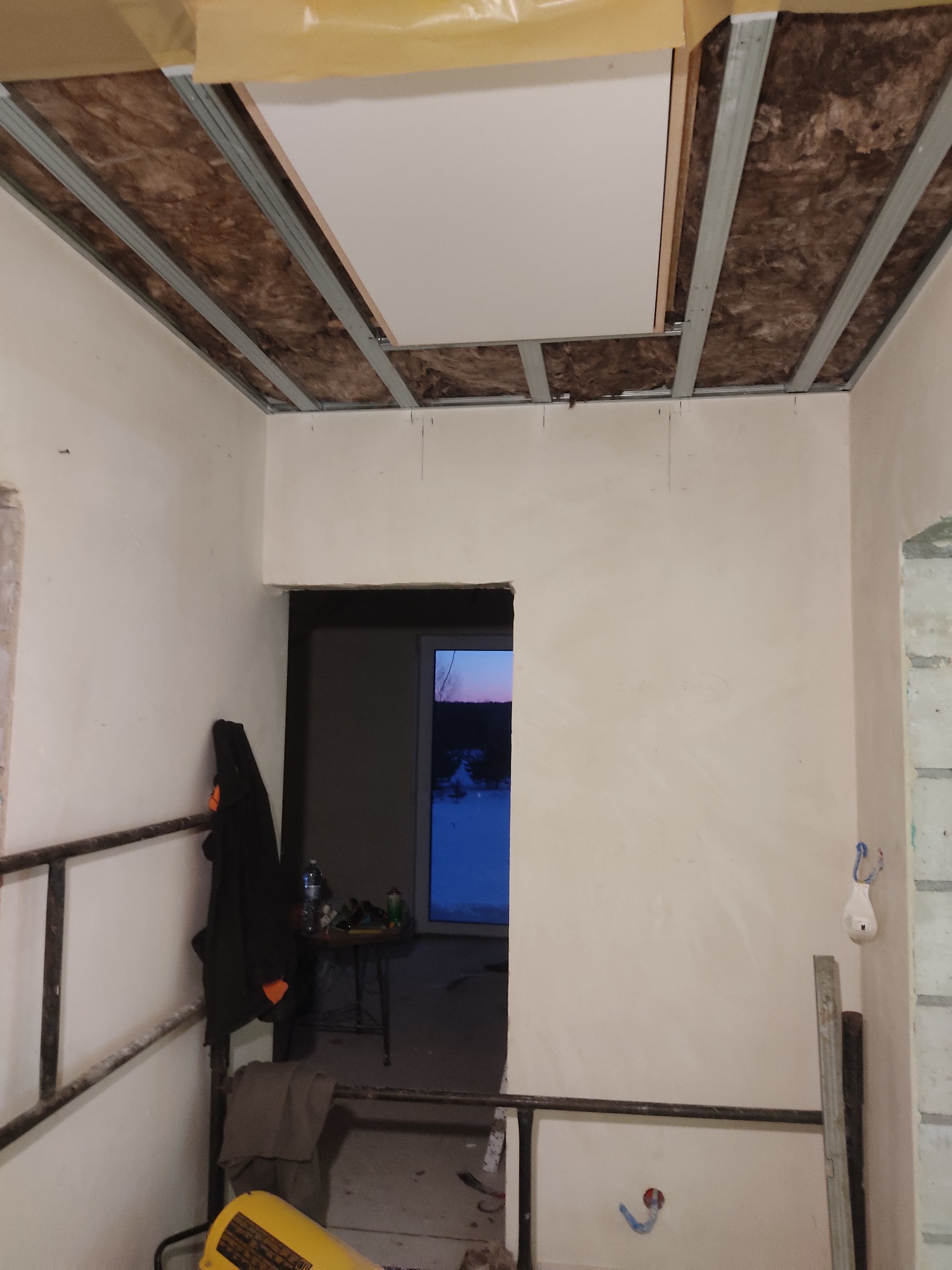 Wnętrze w trakcie remontu: widoczne metalowe profile sufitowe z izolacją z wełny mineralnej, częściowo zamontowana płyta sufitowa, ściany w trakcie szpachlowania, widok na okno i sprzęt budowlany...