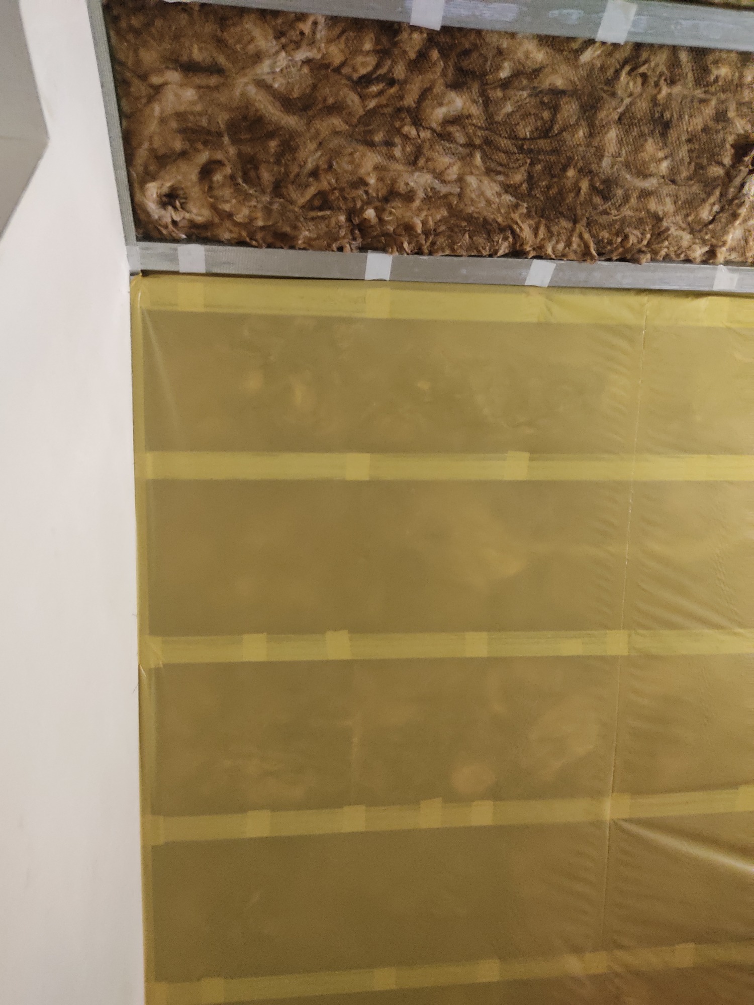 Izolacja z wełny mineralnej pod sufitem, zabezpieczona folią i taśmą malarską na ścianie podczas prac wykończeniowych.