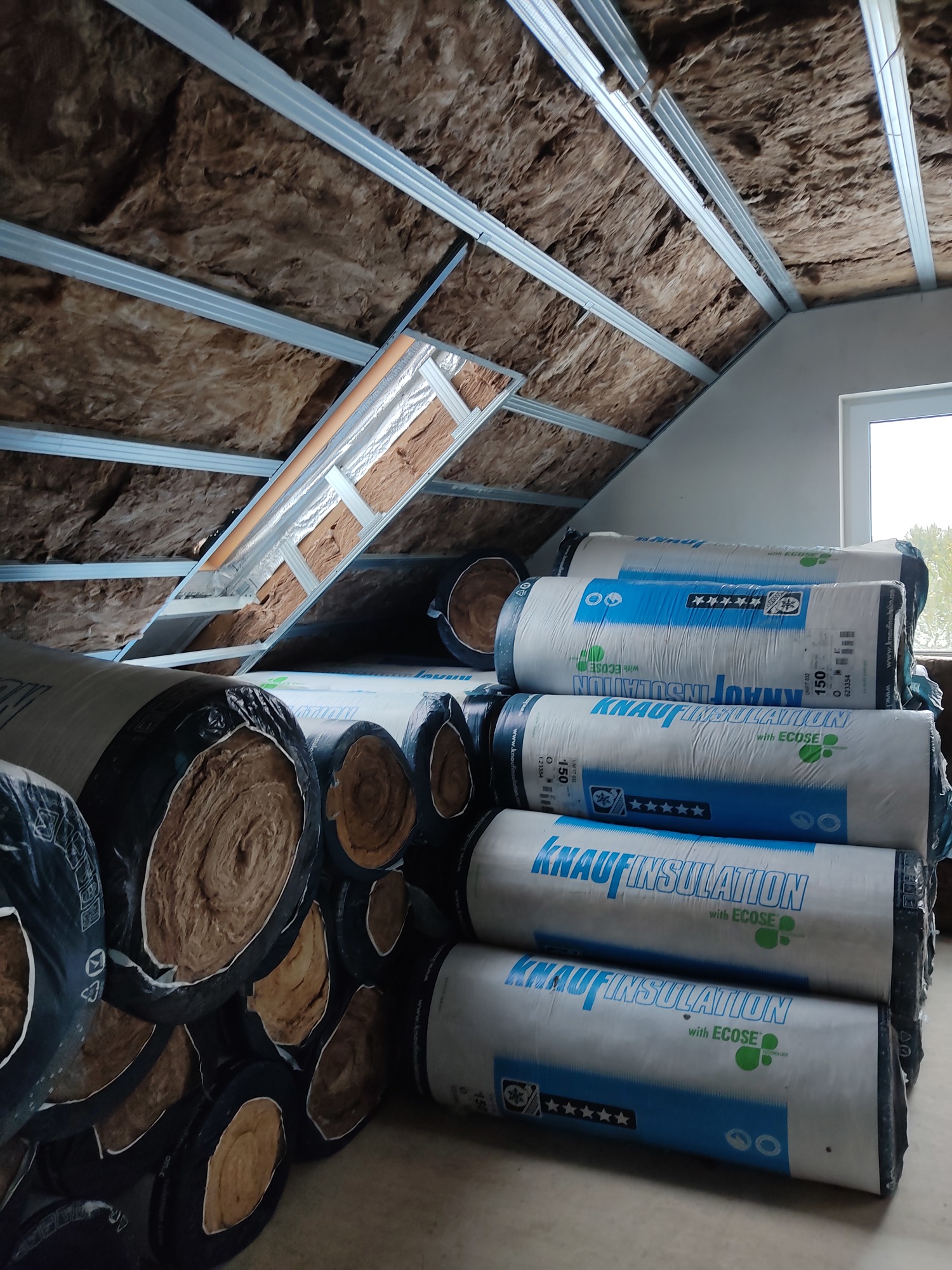 Poddasze w trakcie izolacji: widoczne metalowe profile sufitowe, wełna mineralna pomiędzy nimi oraz rolki wełny Knauf Insulation z etykietą ECOSE ułożone na podłodze.