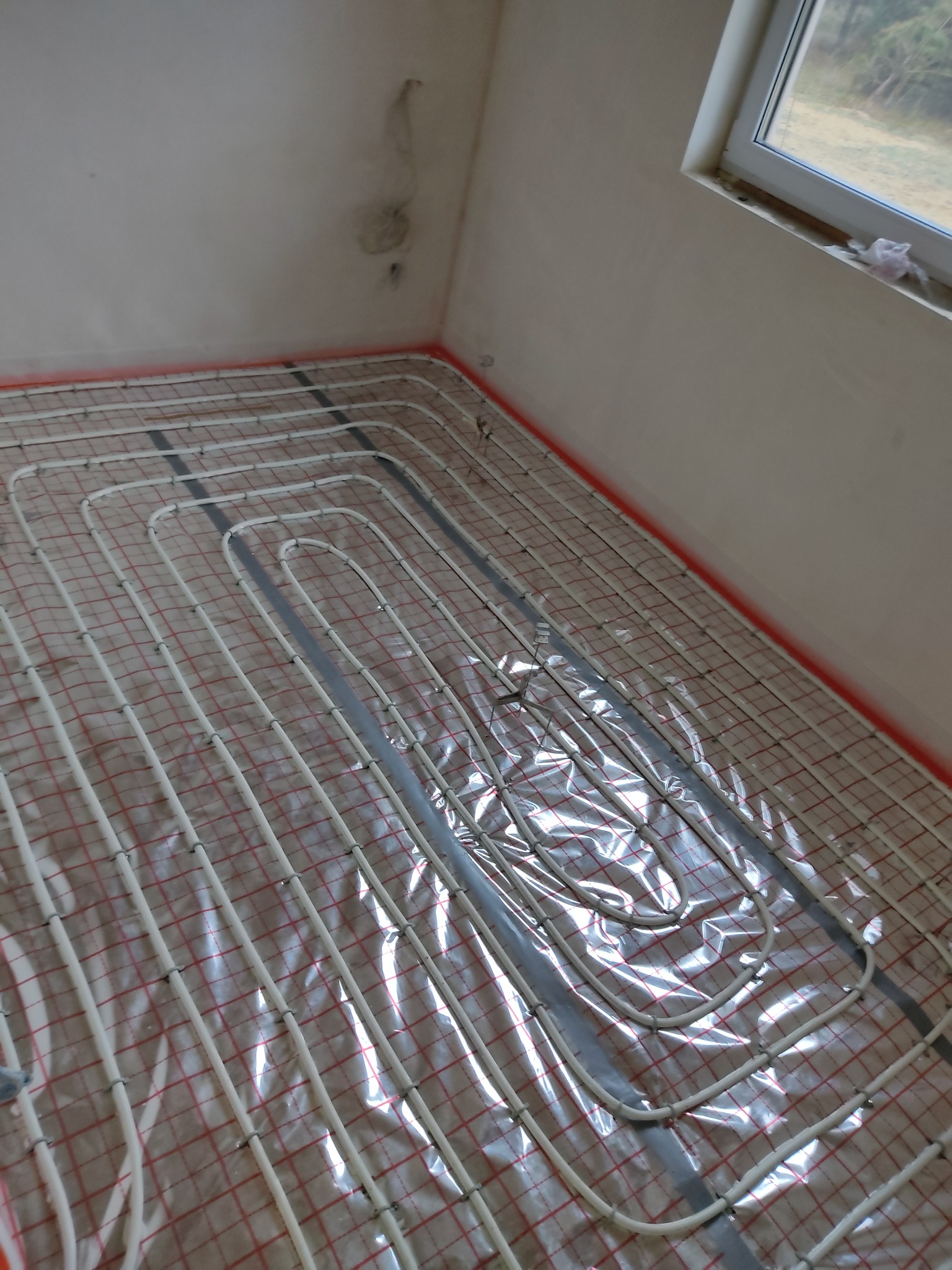 Białe rurki ogrzewania podłogowego ułożone na folii z czerwoną siatką w surowym pomieszczeniu, widok z góry pod kątem.