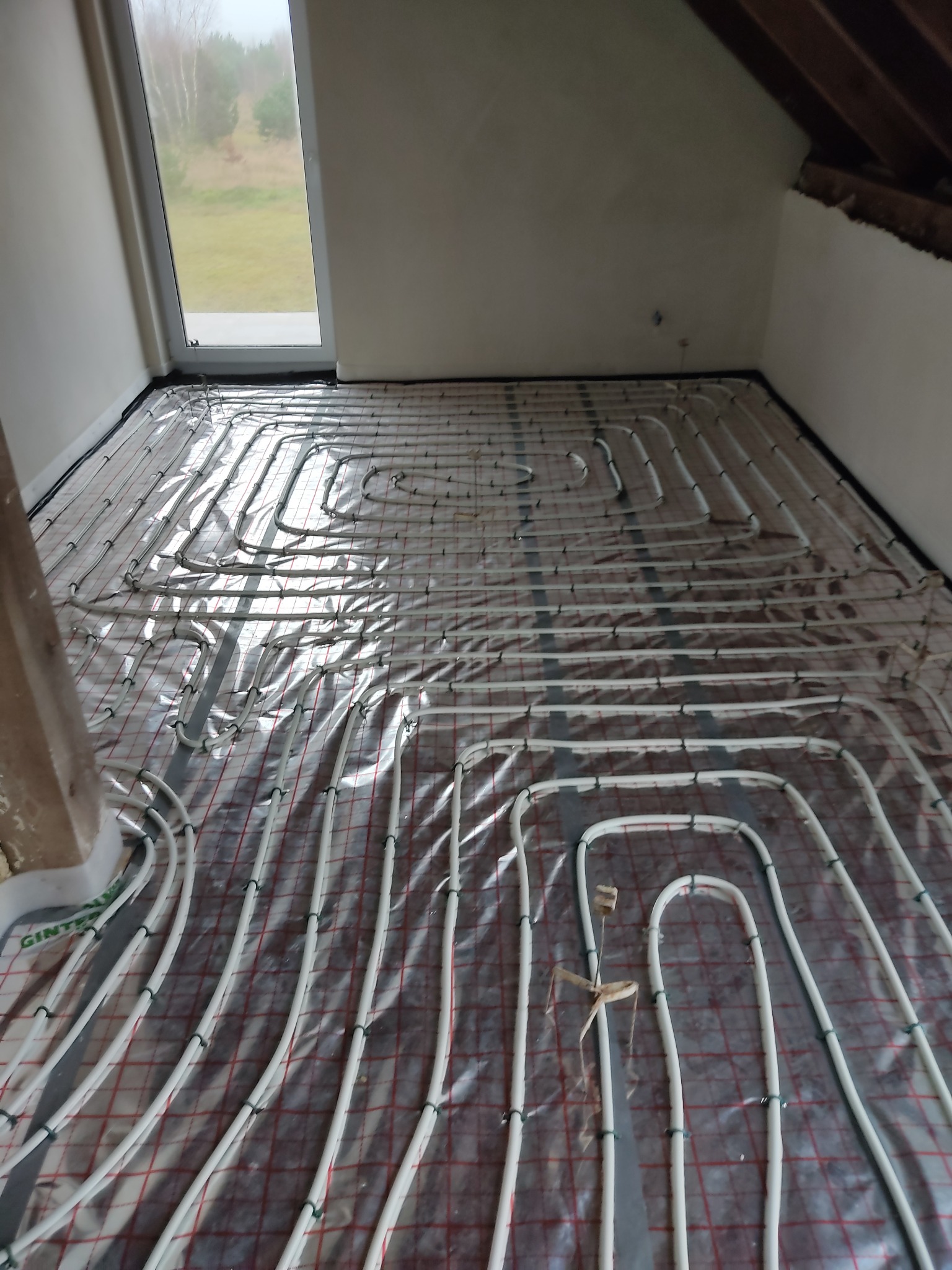 Białe rurki ogrzewania podłogowego ułożone na folii z czerwoną kratką w jasnym pomieszczeniu z oknem, widoczne elementy konstrukcji dachu.