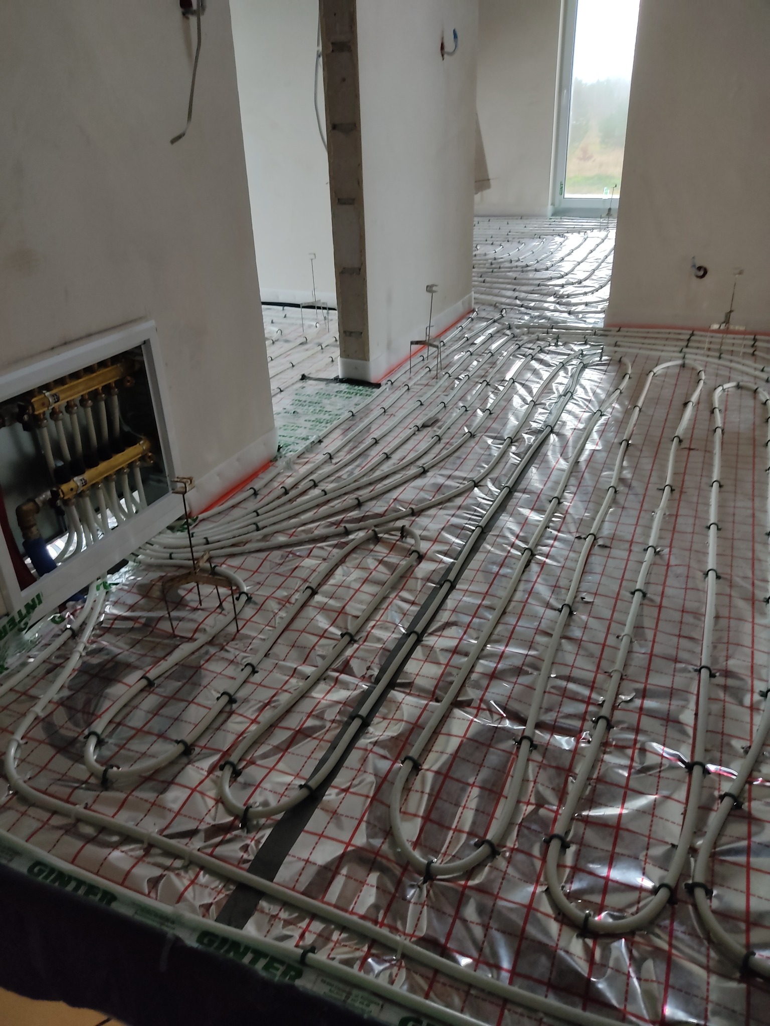 Rozłożone rury ogrzewania podłogowego na folii z siatką w surowym pomieszczeniu, widoczny rozdzielacz z zaworami i okno w tle.