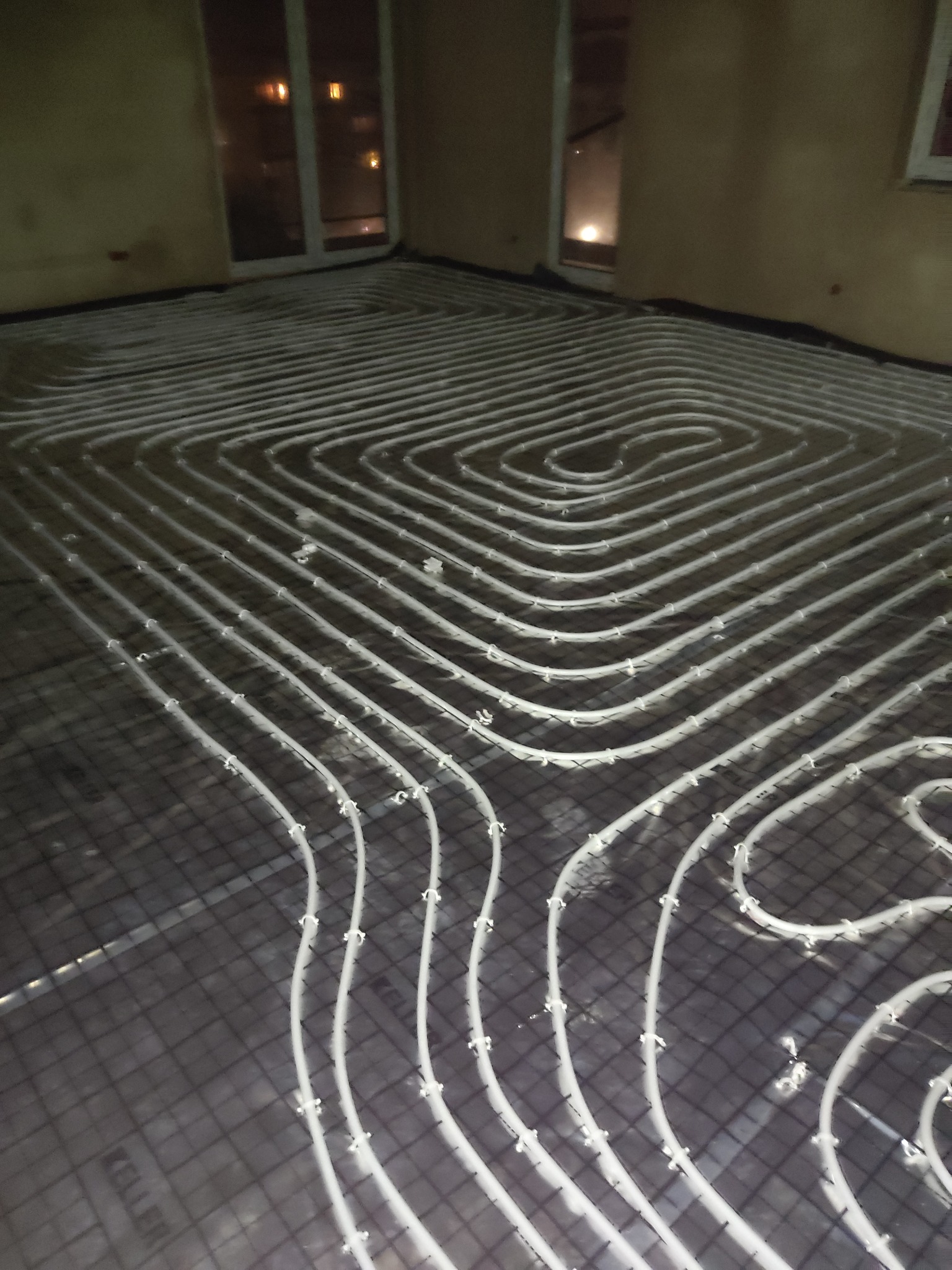 Rozłożone rury ogrzewania podłogowego na siatce, widok z góry, przygotowanie do wylewki jastrychowej, widoczne okno w tle.