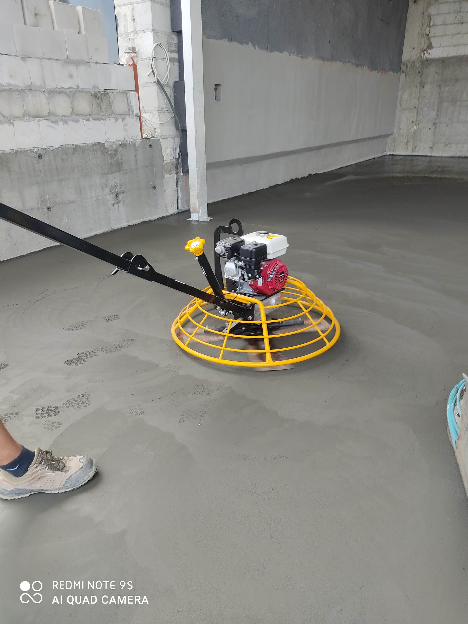 Zacieraczka mechaniczna z żółtym koszem wyrównuje świeżo wylaną, szarą posadzkę w surowym wnętrzu budynku z widocznymi bloczkami betonowymi na ścianach.