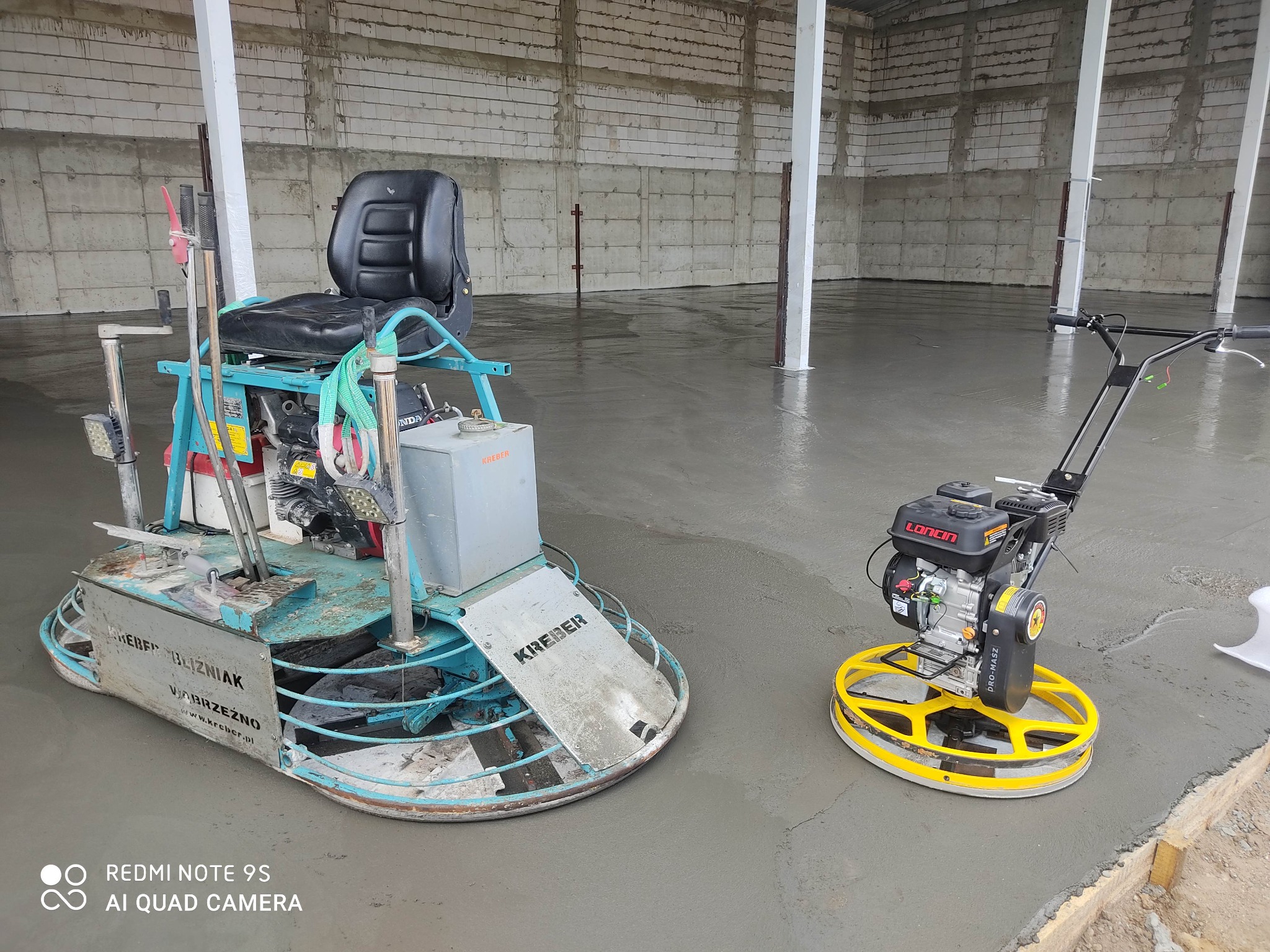 Dwie zacieraczki do betonu, jedna z siedziskiem, druga ręczna, stoją na świeżo wylanej, gładkiej posadzce w surowym wnętrzu hali.