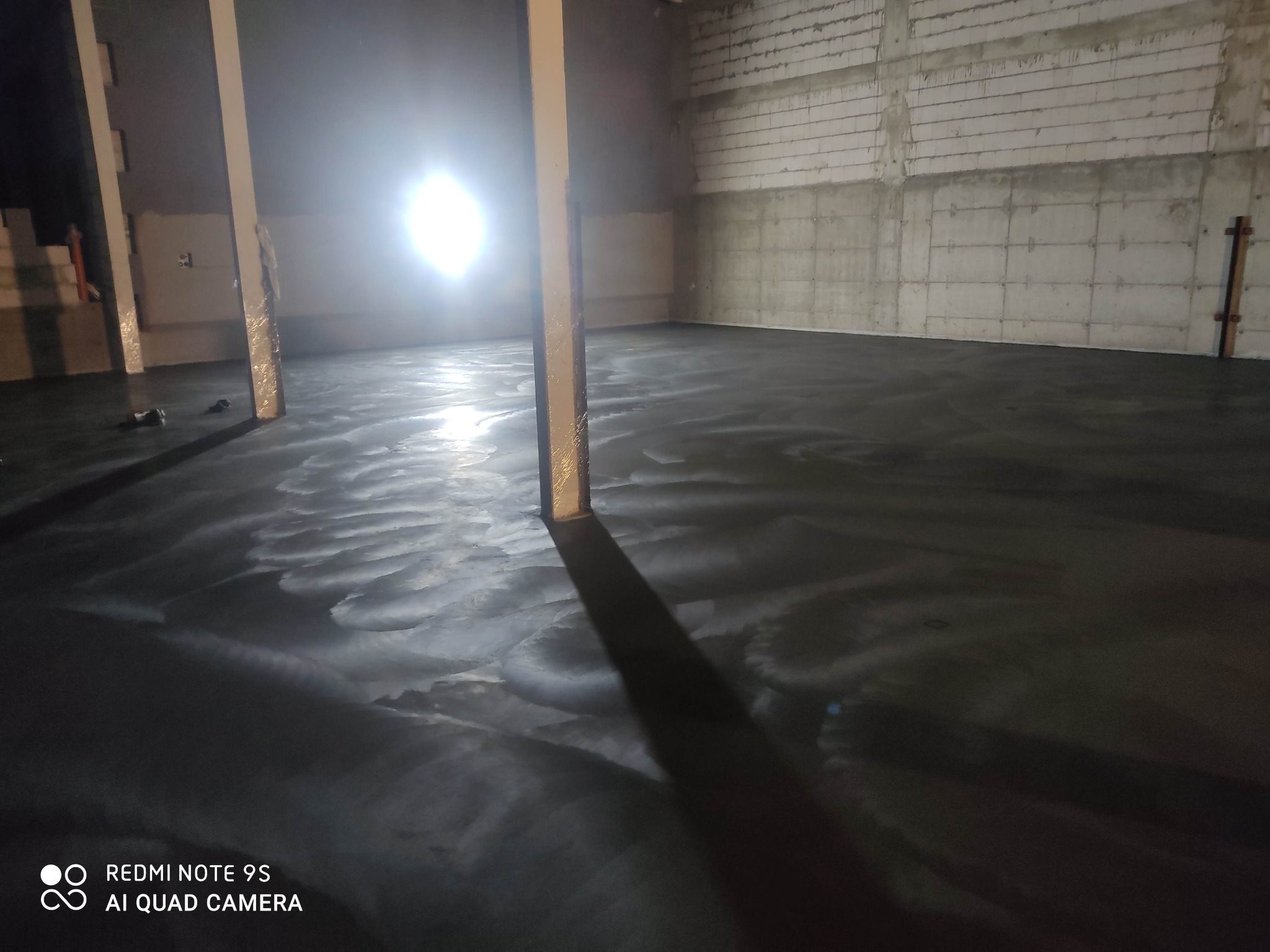 Świeżo wylana, ciemna posadzka w surowym wnętrzu z widocznymi śladami rozprowadzania, oświetlona mocnym światłem punktowym, z surowymi ścianami z betonu komórkowego.