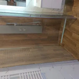 Szklana kabina prysznicowa z panelem natryskowym zamontowana w narożniku łazienki wyłożonej płytkami imitującymi drewno, widoczny grzejnik drabinkowy.