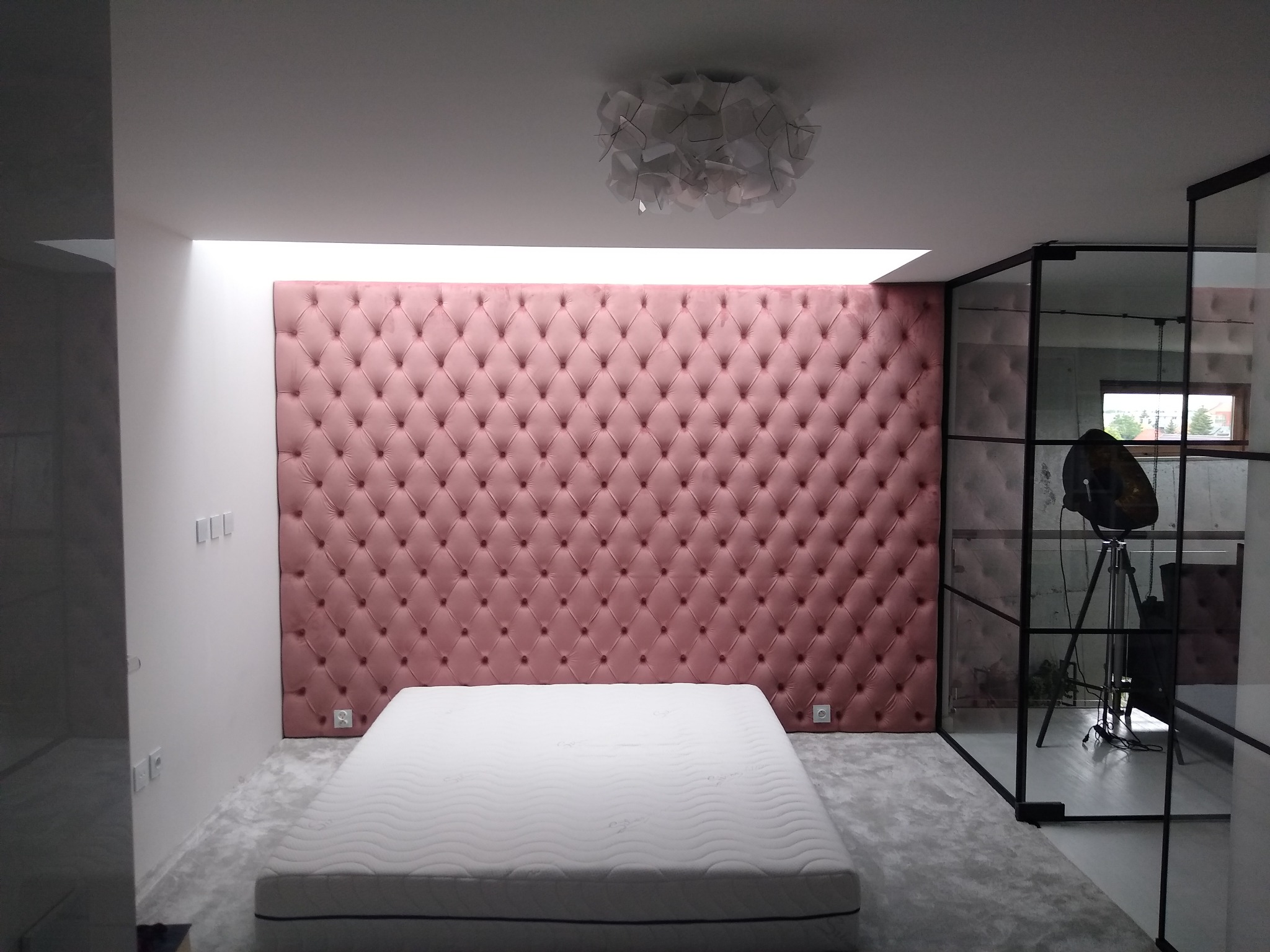 Różowy, pikowany panel ścienny za materacem w minimalistycznej sypialni z szarą wykładziną i szklaną ścianą działową, oświetlony górnym światłem i nowoczesną lampą sufitową.