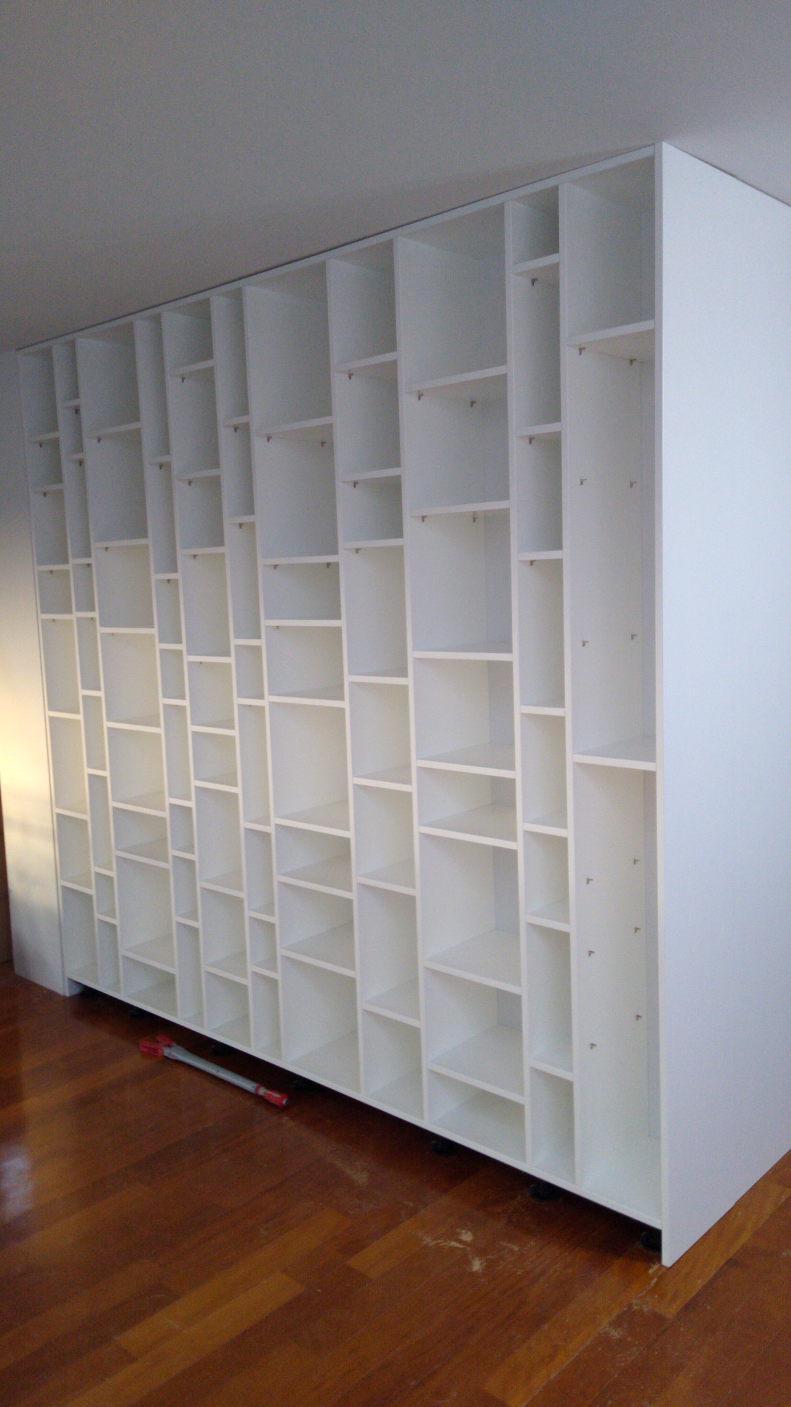 Biała, modułowa konstrukcja regałowa z licznymi półkami o różnej wielkości, zamontowana pod sufitem, widoczna na tle drewnianej podłogi i jasnej ściany, z widocznym narzędziem do montażu leżącym...