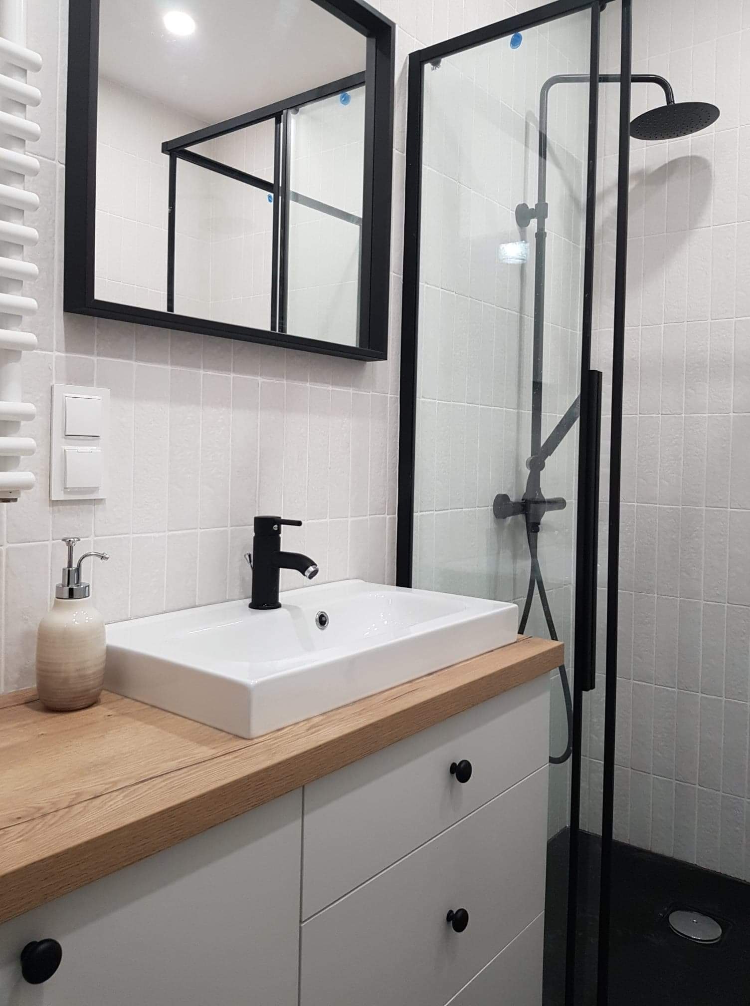 Nowoczesna łazienka z drewnianym blatem, białą umywalką i czarną armaturą, kabina prysznicowa z czarną ramą i deszczownicą, lustro w czarnej ramie.