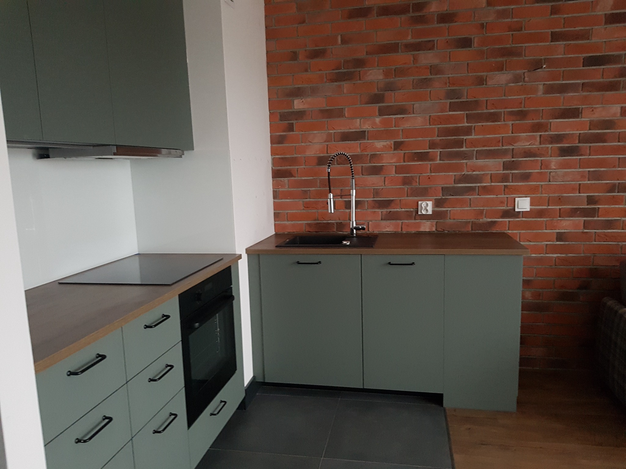 Fragment szarej kuchni z ceglaną ścianą w tle, widoczny zlew z elastyczną wylewką, płyta indukcyjna i piekarnik.