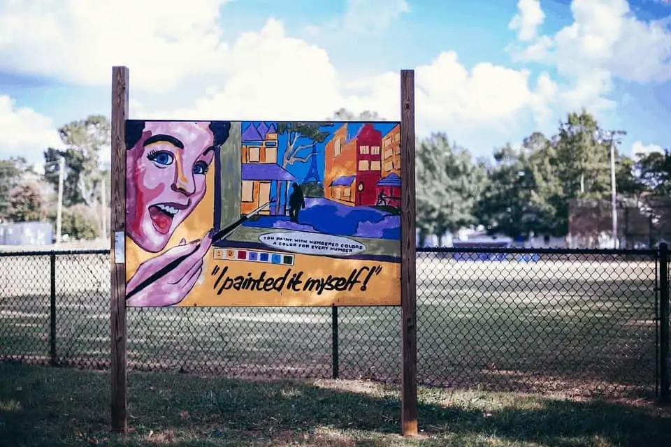 Duży plakat z kolorową ilustracją przedstawiającą kobietę malującą obraz z wieżowcami i wieżą Eiffla, umieszczony na drewnianych słupkach na tle trawnika i ogrodzenia.