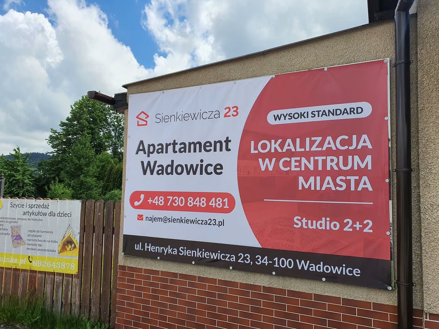 Baner reklamowy apartamentów w Wadowicach, z widocznym adresem, numerem telefonu i adresem e-mail, zawieszony na ścianie budynku.