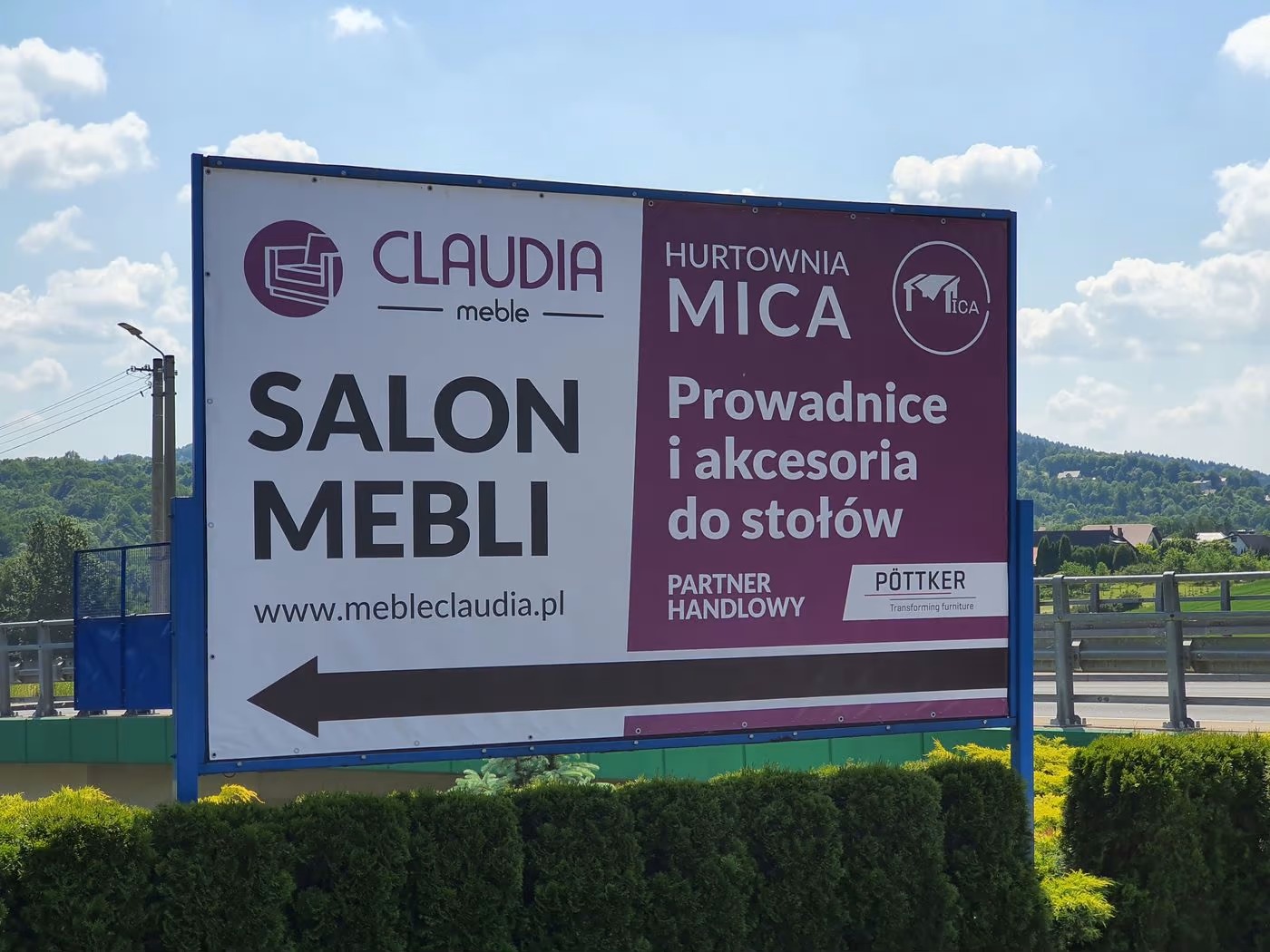 Billboard reklamowy salonu meblowego Claudia i hurtowni Mica z informacją o prowadnicach i akcesoriach do stołów, widoczny przy drodze na tle zieleni i błękitnego nieba.