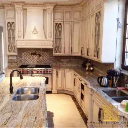 Jasna kuchnia w stylu amerykańskim z wyspą kuchenną, marmurowym blatem, białymi szafkami z ozdobnymi przeszkleniami i okapem z ornamentem. Widoczny fragment podłogi i okno.