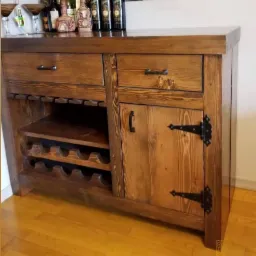 Rustykalny barek sosnowy z metalowymi okuciami, widoczne dwie szuflady, półki na butelki i szafka z ozdobnymi zawiasami, na blacie barku ustawione butelki i kieliszki.