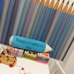 Pokój dziecięcy z tapetą w niebieskie kredki, poduszką w kształcie kredki z napisem 'Pracownia Pedagogiczno-Logopedyczna Blue', drewnianą zabawką edukacyjną i stosem książeczek na białym stoliku.