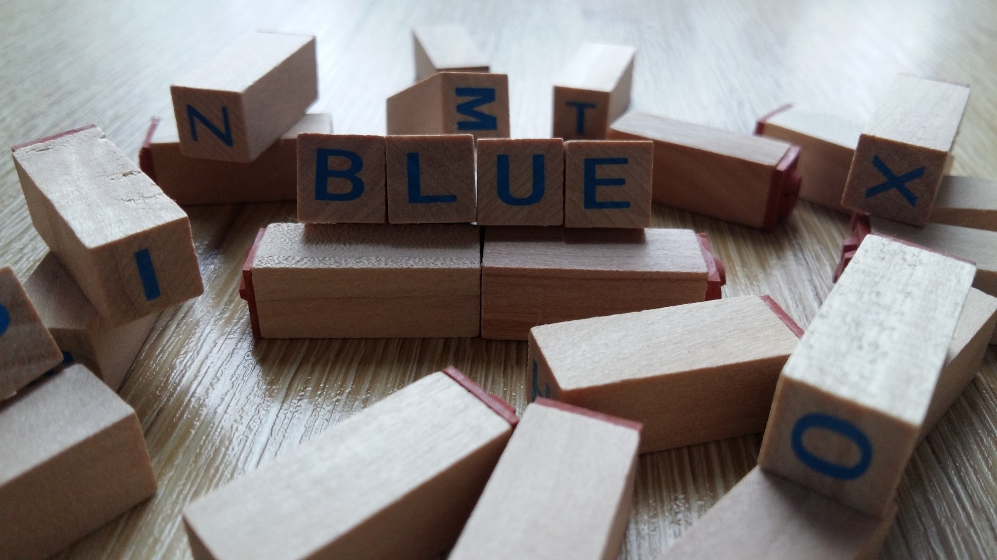 Drewniane klocki z literami układające się w słowo 'BLUE' na drewnianej powierzchni, otoczone innymi klockami z pojedynczymi literami.