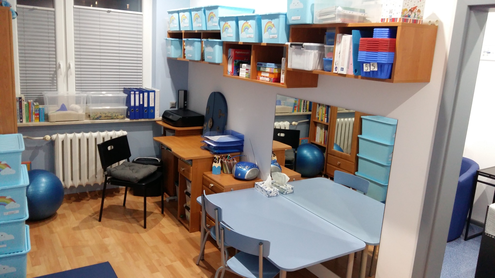 Gabinet logopedyczny z biurkiem, stolikiem z lustrem, regałami z książkami i pojemnikami na zabawki, utrzymany w jasnej kolorystyce.