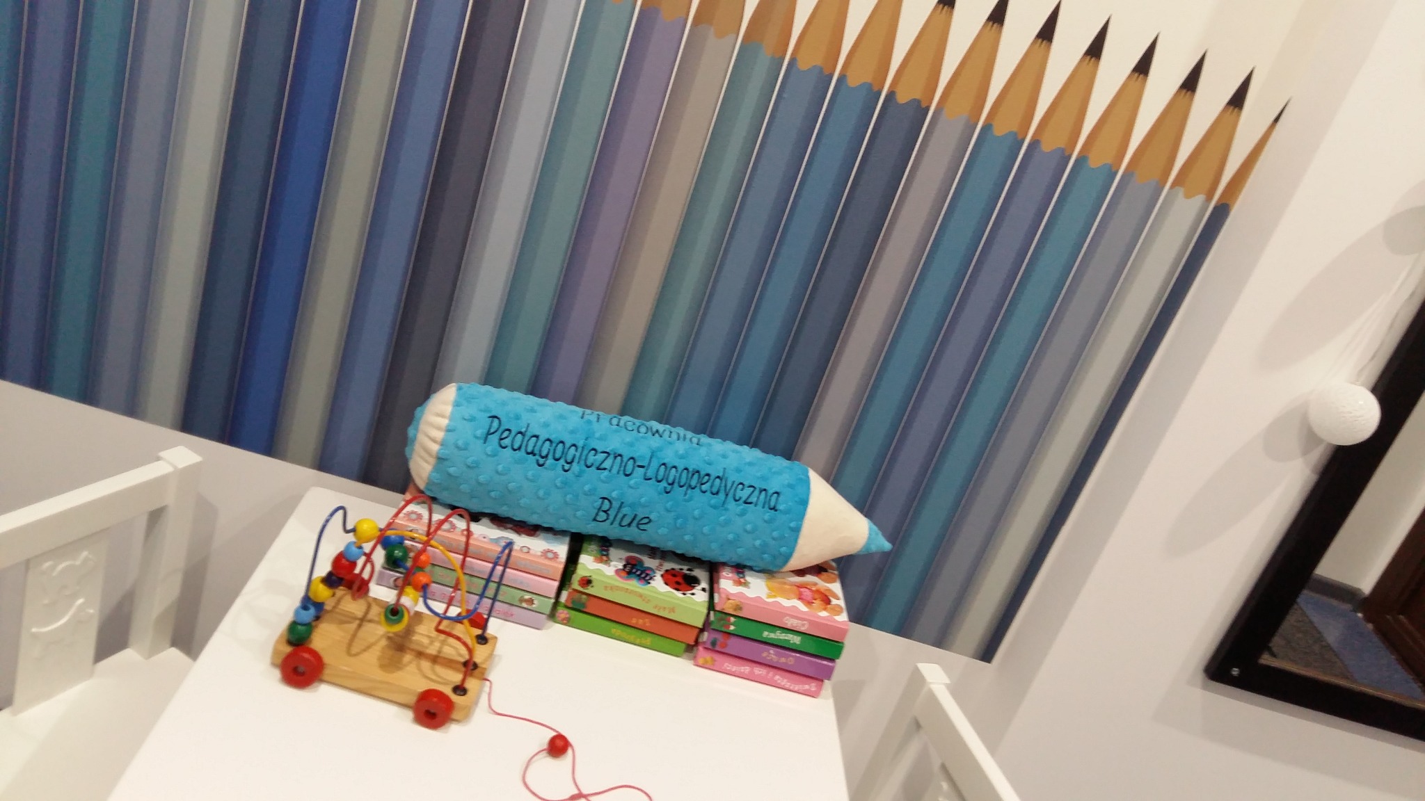 Pokój dziecięcy z tapetą w niebieskie kredki, poduszką w kształcie kredki z napisem 'Pracownia Pedagogiczno-Logopedyczna Blue', drewnianą zabawką edukacyjną i stosem książeczek na białym stoliku.