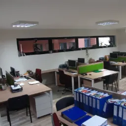 Widok przestronnego biura z biurkami, komputerami, segregatorami i oknem z widokiem na budynek z czerwonej cegły.