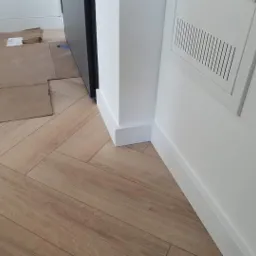 Ułożona pod kątem drewnopodobna podłoga, zabezpieczona kartonem przy ścianie z białą listwą przypodłogową i skrzynką wentylacyjną.