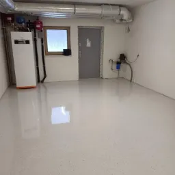 Jasnoszara, błyszcząca posadzka z drobnymi, ciemnymi plamkami w pomieszczeniu technicznym z kotłem grzewczym, oknem i szarą metalową bramą.