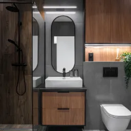 Nowoczesna łazienka z szarymi płytkami, drewnianymi akcentami, owalnym lustrem, wiszącą toaletą i czarną armaturą, oświetlona listwą LED pod szafką.