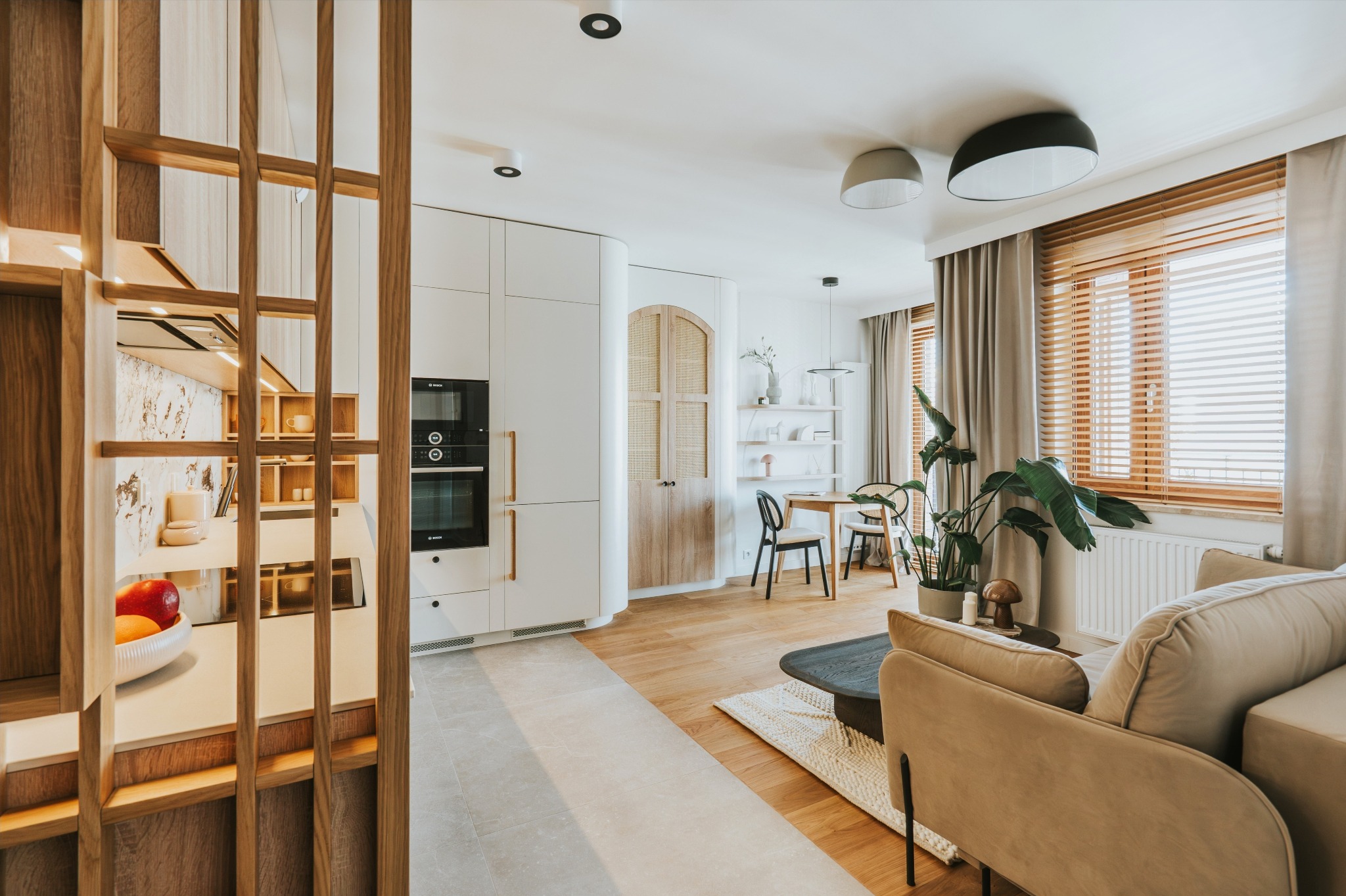 Jasne, przestronne wnętrze z drewnianą ażurową ścianką działową, nowoczesną zabudową kuchenną i strefą relaksu z beżową sofą. Parkiet w jodełkę.