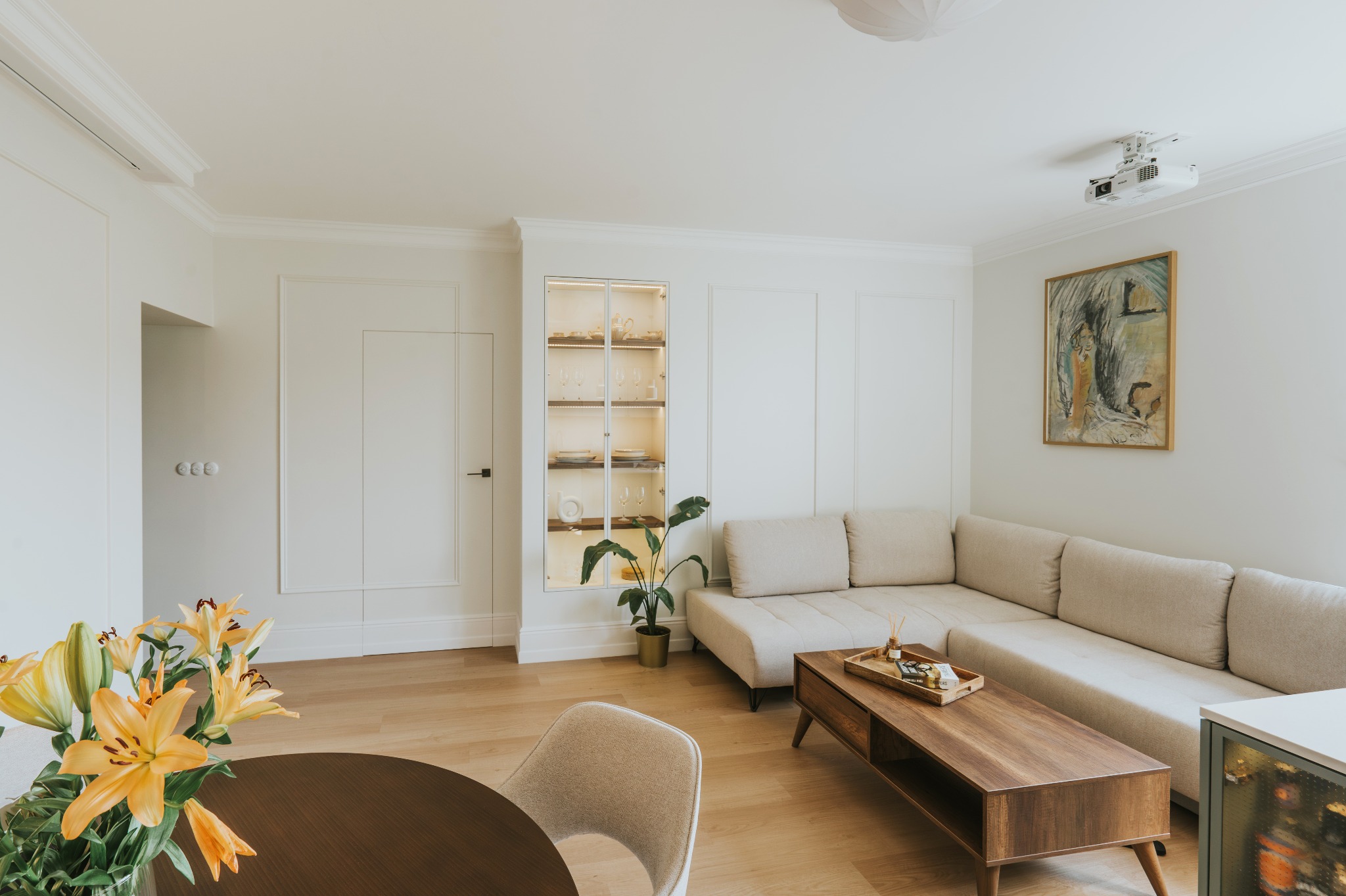 Jasne wnętrze salonu z narożną sofą, drewnianym stolikiem kawowym i okrągłym stołem. Na ścianie wbudowana witryna z oświetleniem i obraz. Projektor na suficie.