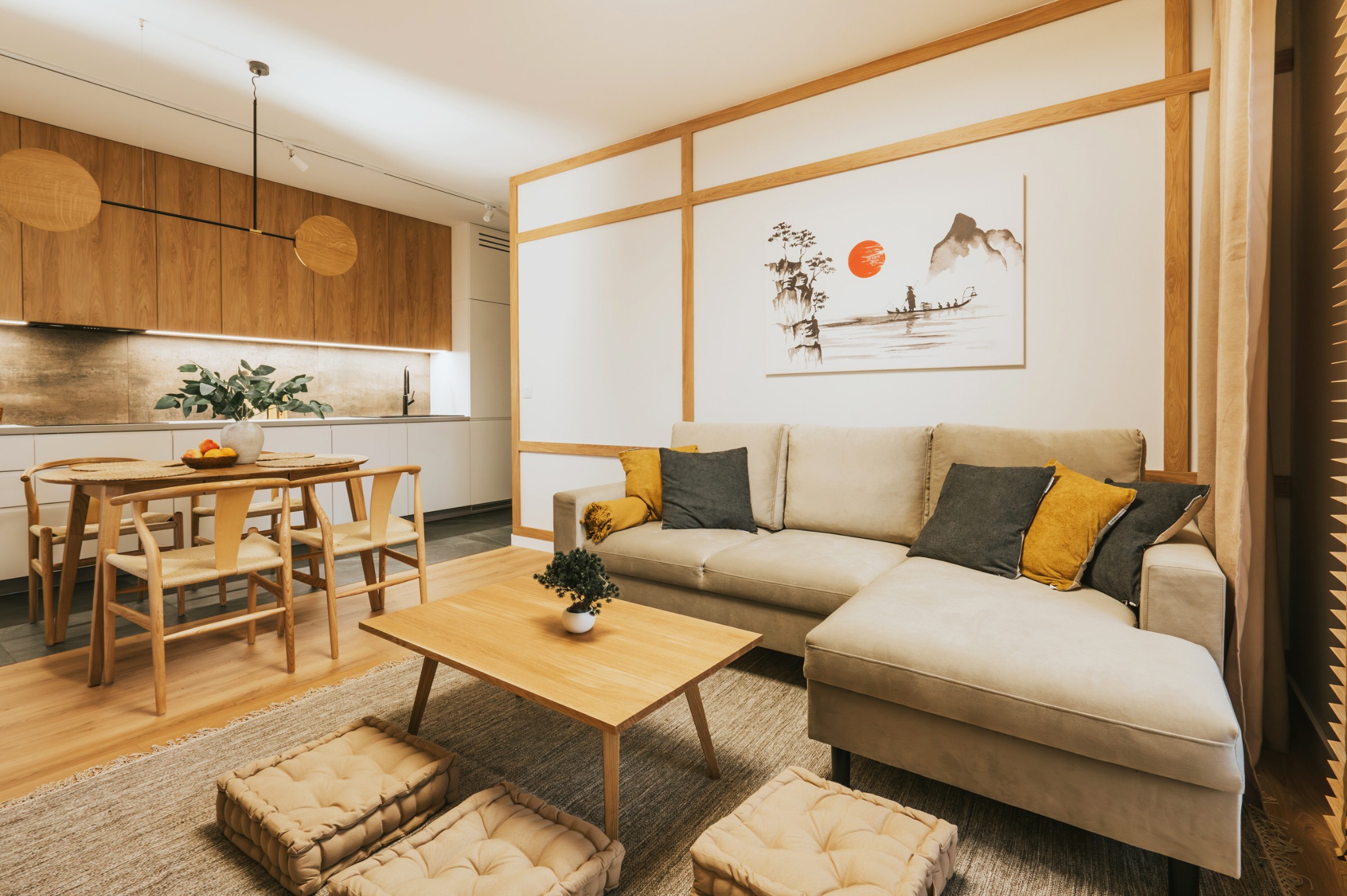 Jasne, przestronne wnętrze mieszkania z drewnianymi akcentami: stół z krzesłami, sofa z poduszkami, dywan z pufami, obraz w stylu japońskim. Oświetlenie punktowe i lampa wisząca.