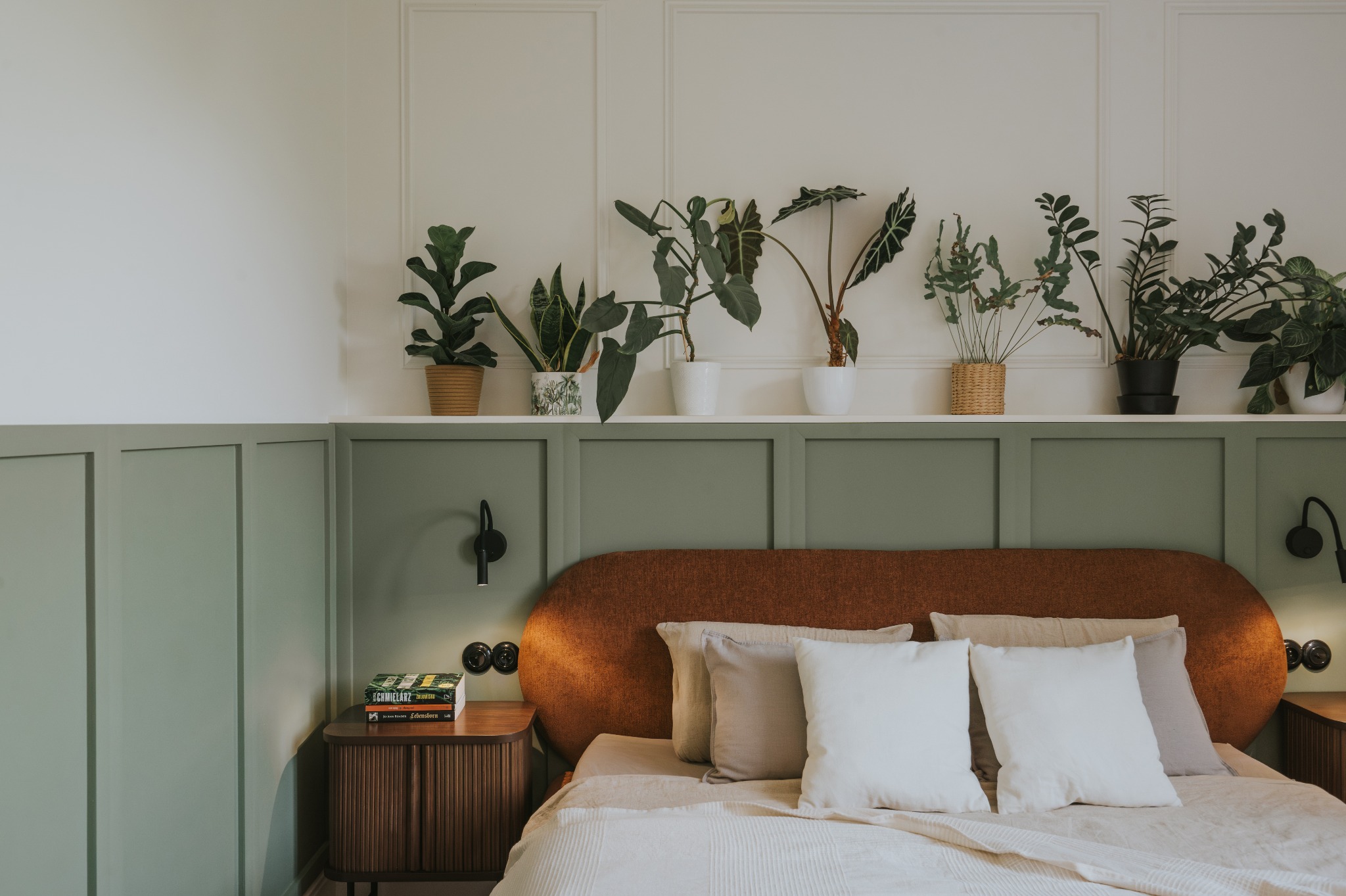 Stylowa sypialnia z zielonymi panelami ściennymi, roślinami na półce i drewnianą szafką nocną. Uwagę przykuwa pomarańczowe wezgłowie łóżka i lampki ścienne.