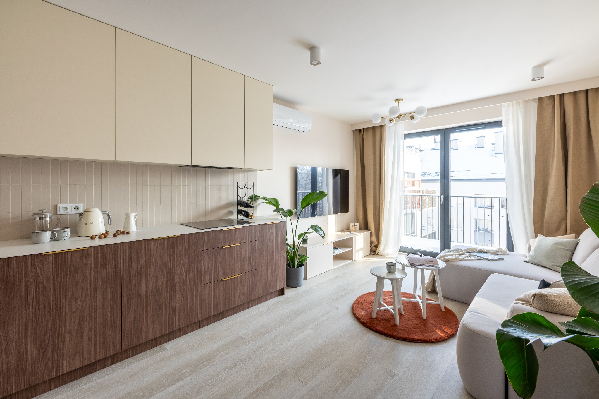 Jasne wnętrze mieszkania z drewnianą zabudową kuchenną, beżowymi szafkami i balkonem z widokiem na miasto, oświetlone naturalnym światłem.