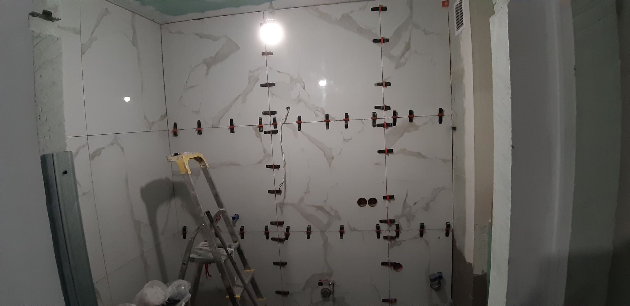 Ściana w trakcie układania płytek imitujących marmur, z użyciem poziomicy laserowej i klipsów dystansowych, widoczna drabina malarska.