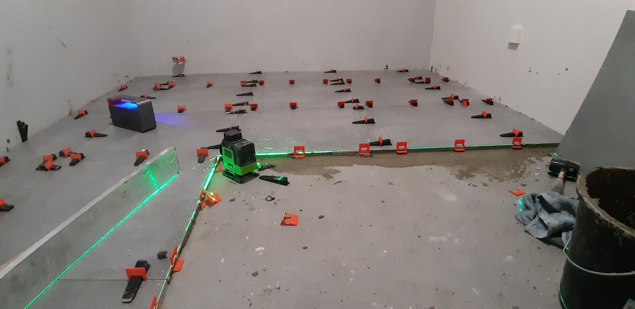 Przygotowanie do układania płytek podłogowych: poziomowanie laserowe, klipsy dystansowe i kliny do wyrównywania płytek, zaprawa w wiadrze, młotek gumowy.