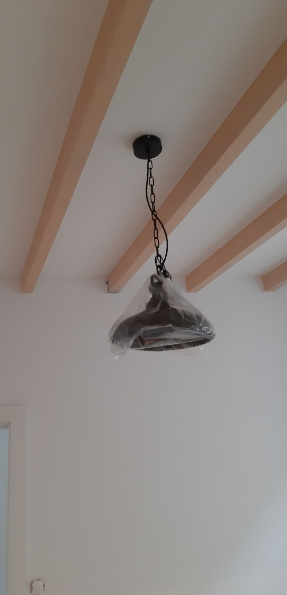 Lampa wisząca w folii ochronnej, zamontowana na suficie z widocznymi drewnianymi belkami dekoracyjnymi.