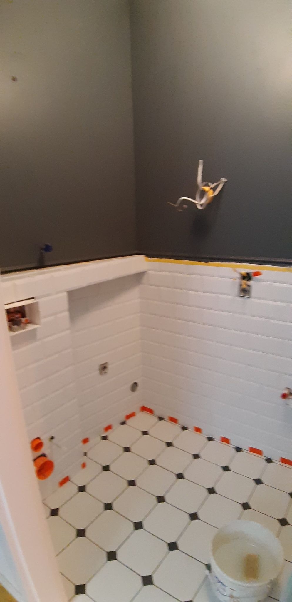 Łazienka w trakcie remontu: białe, poziome płytki ścienne z fugą, czarno-biała podłoga w romby z pomarańczowymi krzyżykami dystansowymi, instalacja elektryczna wystająca ze ściany, szara ściana...