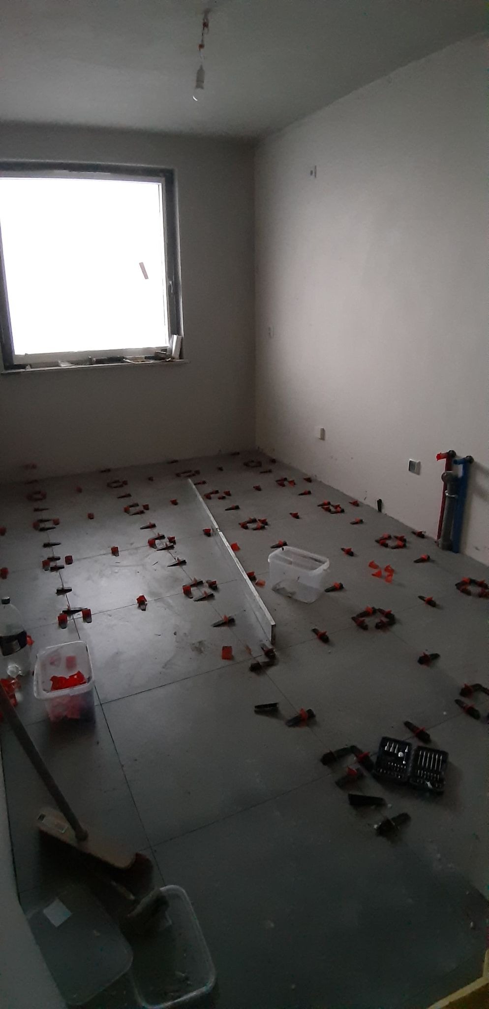 Szare płytki podłogowe w trakcie układania, widoczne systemy poziomowania płytek, puste wiaderko i narzędzia na podłodze w słabo oświetlonym pomieszczeniu z oknem.