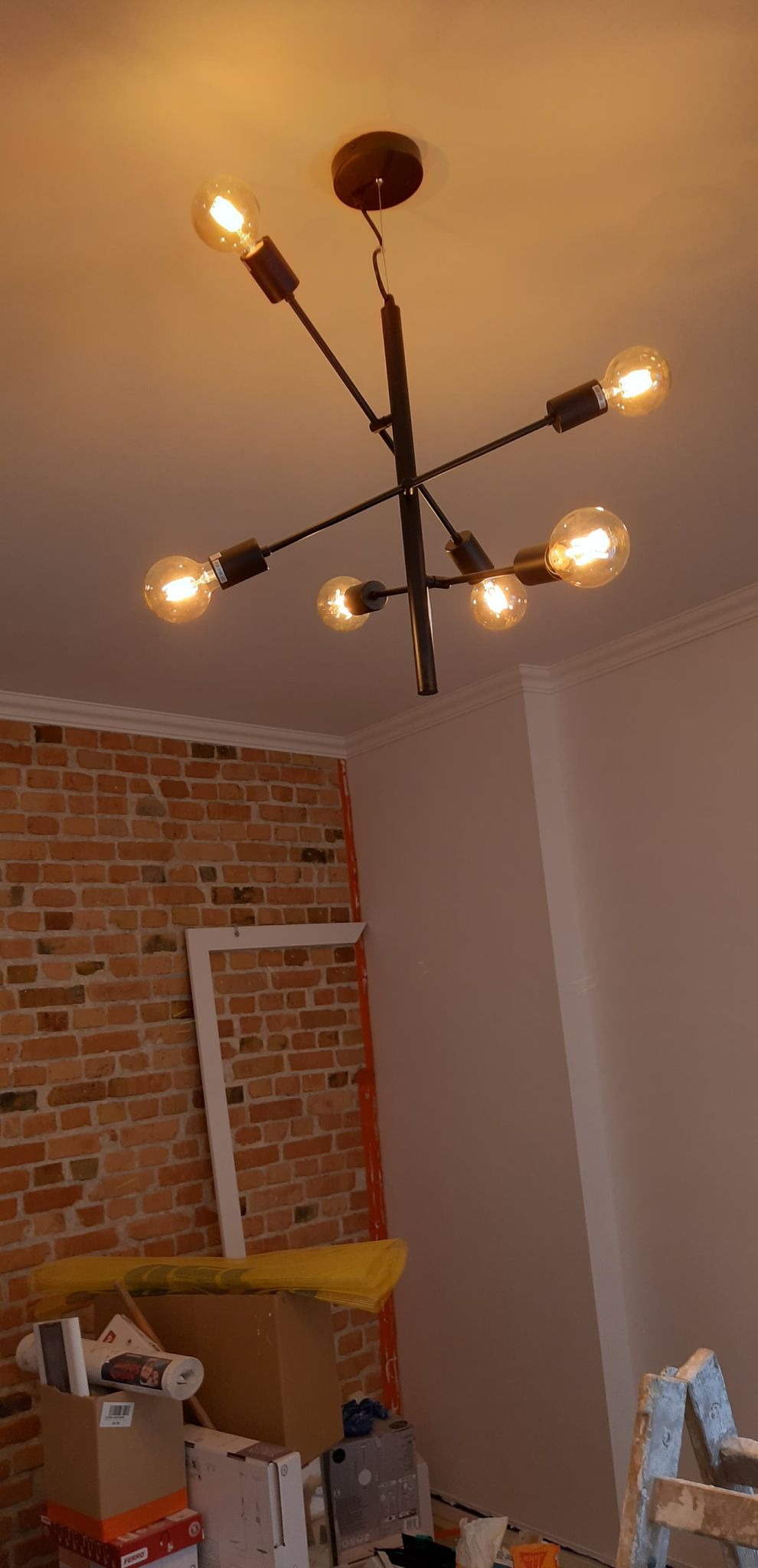 Czarna, nowoczesna lampa sufitowa z sześcioma żarówkami edisona, zamontowana w pomieszczeniu z częściowo odsłoniętą ścianą z czerwonej cegły i białymi ścianami, widoczne przygotowania do prac...