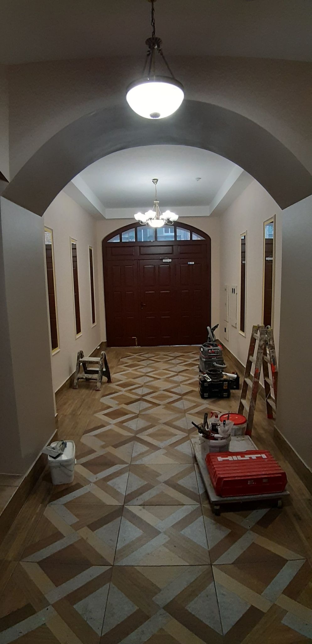 Korytarz z nową, wzorzystą podłogą z płytek, narzędzia glazurnicze i drabina w tle, ściany pomalowane na jasny kolor, widoczne elementy oświetlenia sufitowego.