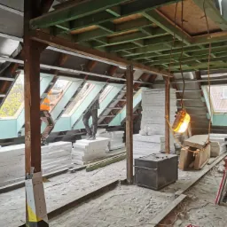 Poddasze w trakcie prac izolacyjnych: pracownicy stoją na ułożonych warstwach izolacji, montaż płyt gipsowo-kartonowych przy oknach dachowych, widoczne elementy konstrukcji drewnianej i oświetlenie...