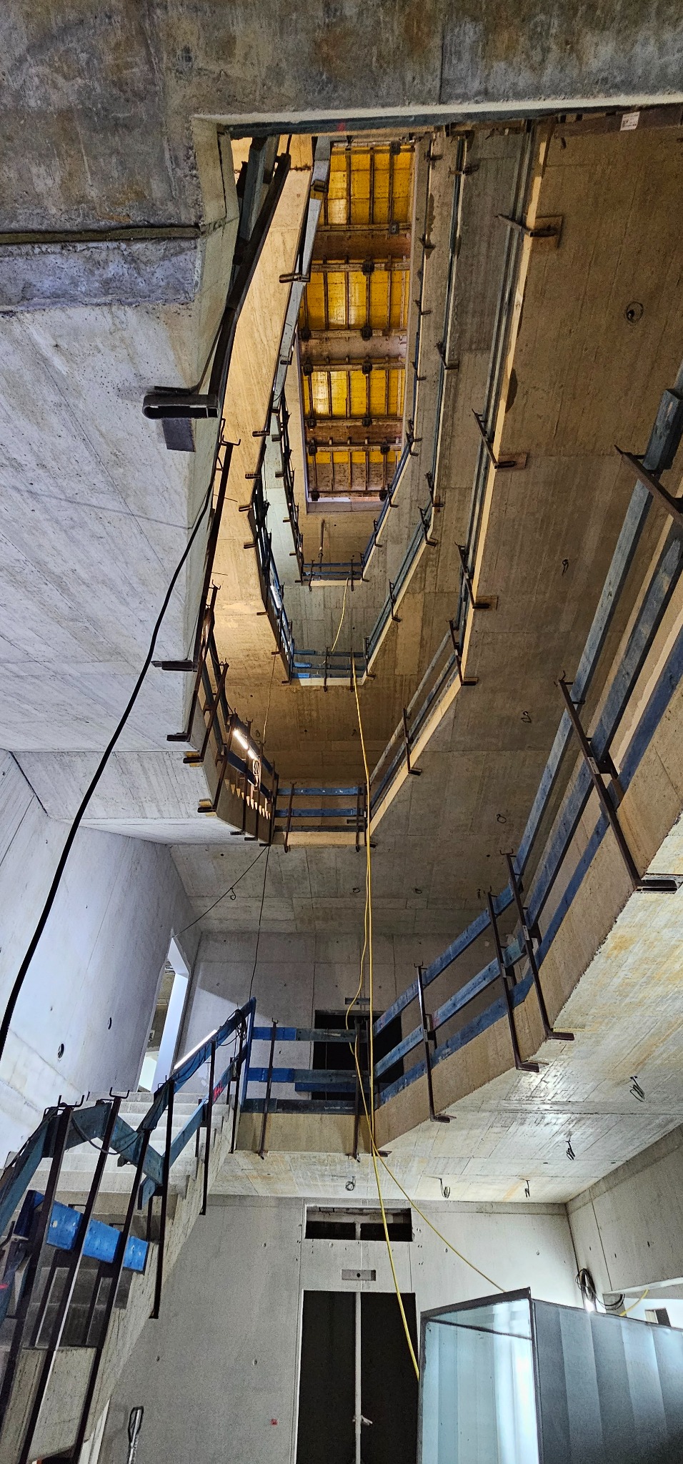 Wnętrze w trakcie budowy: surowe betonowe schody spiralne z drewnianymi barierkami, widok z dołu w górę, żółte szalunki widoczne w otworze stropu, przewody elektryczne zwisające z sufitu, ściany...