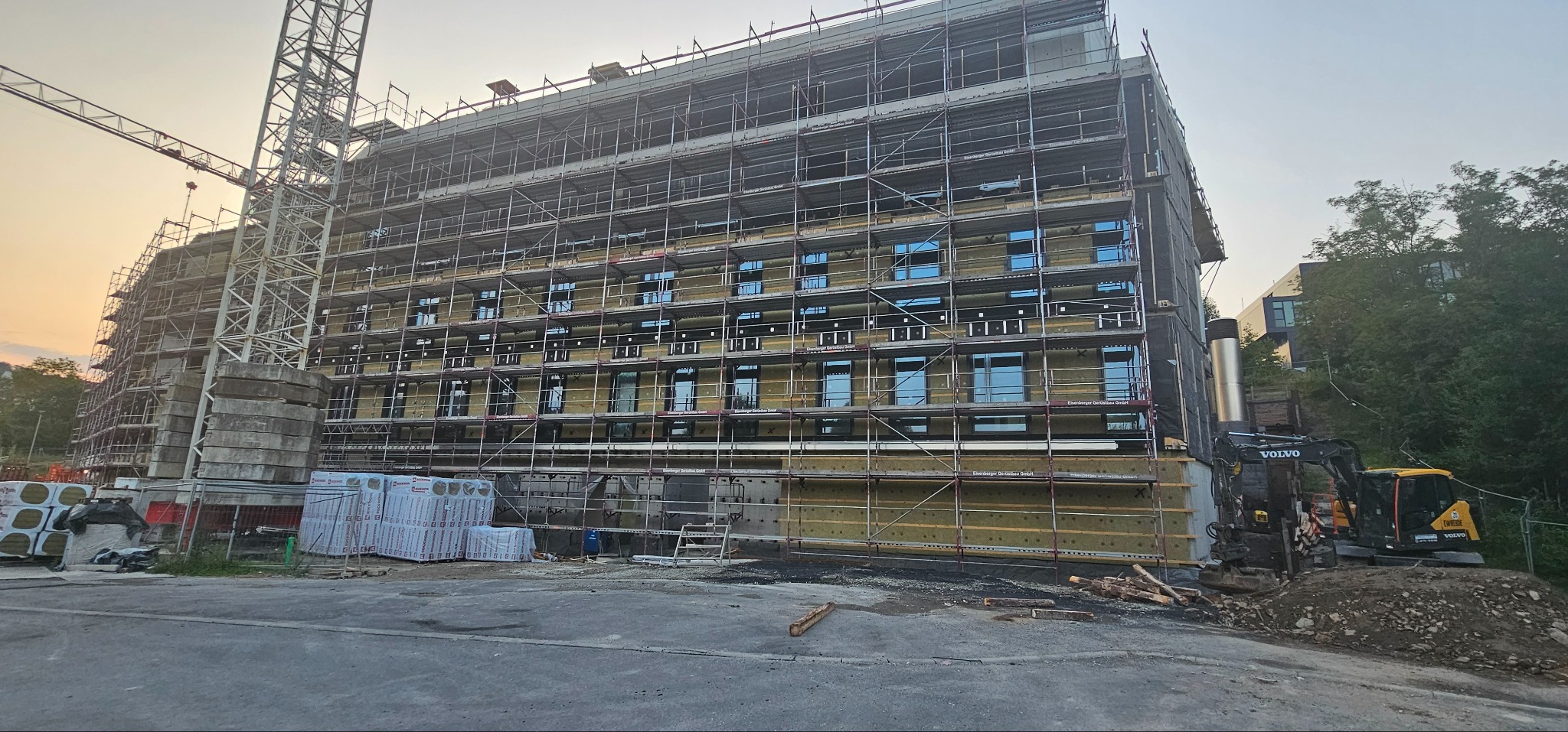 Budynek w trakcie ocieplania elewacji, widoczne rusztowania, materiały izolacyjne i koparka Volvo na placu budowy.