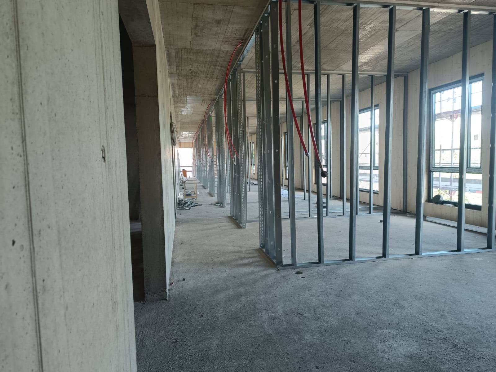 Wnętrze w stanie surowym z metalową konstrukcją ścian działowych i instalacją elektryczną w czerwonych rurach karbowanych, widoczne okna i surowy betonowy strop.