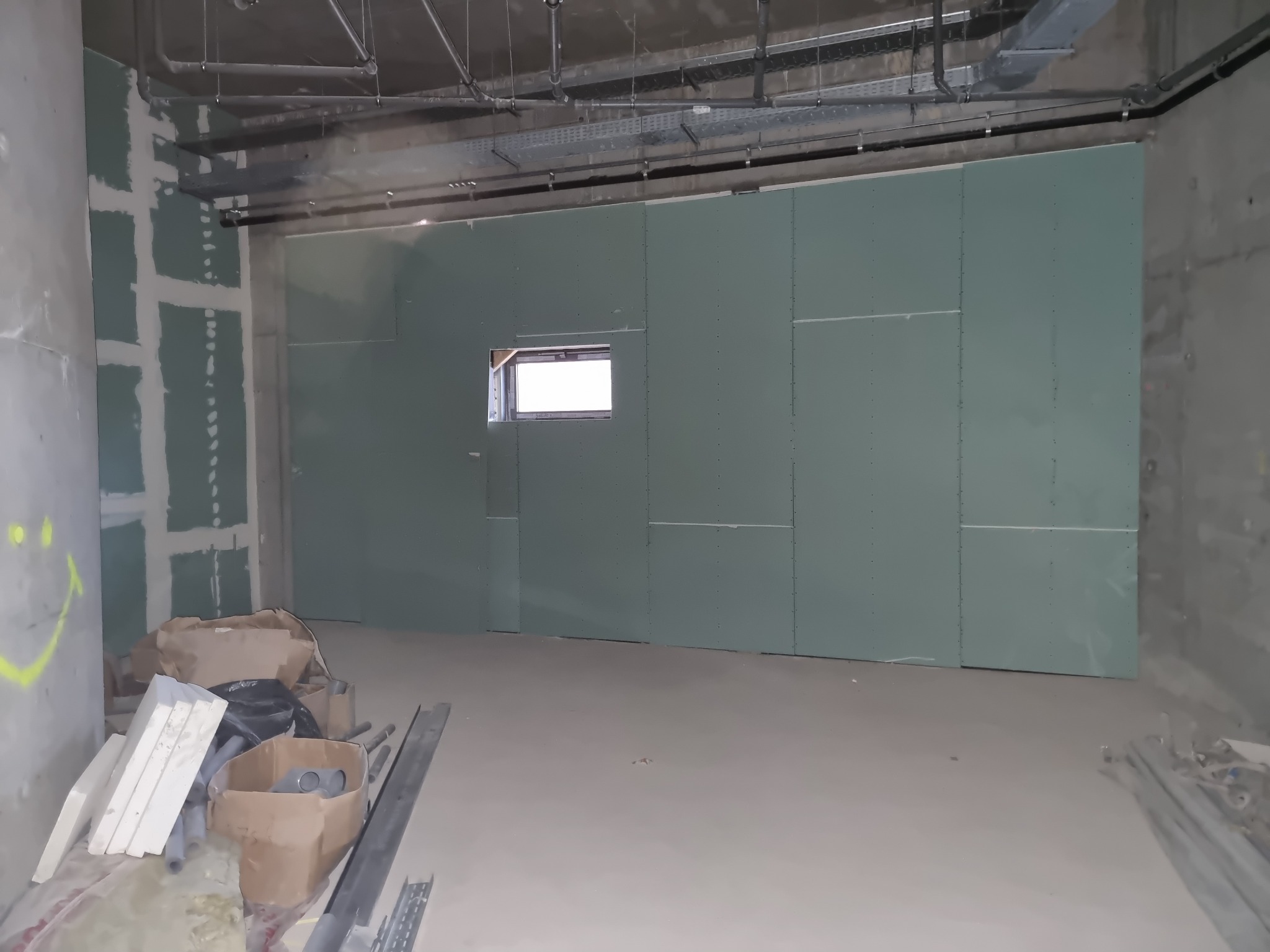 Wnętrze w trakcie prac wykończeniowych: ściana z płyt karton gips, z widocznym otworem okiennym i instalacją pod sufitem. Na ścianie po lewej stronie widoczny żółty, namalowany uśmiech.