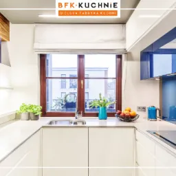 Jasna kuchnia w stylu minimalistycznym z drewnianą ramą okienną, widok na budynek z oknami, na blacie owoce w koszyku, niebieski czajnik i wazon z tulipanami.