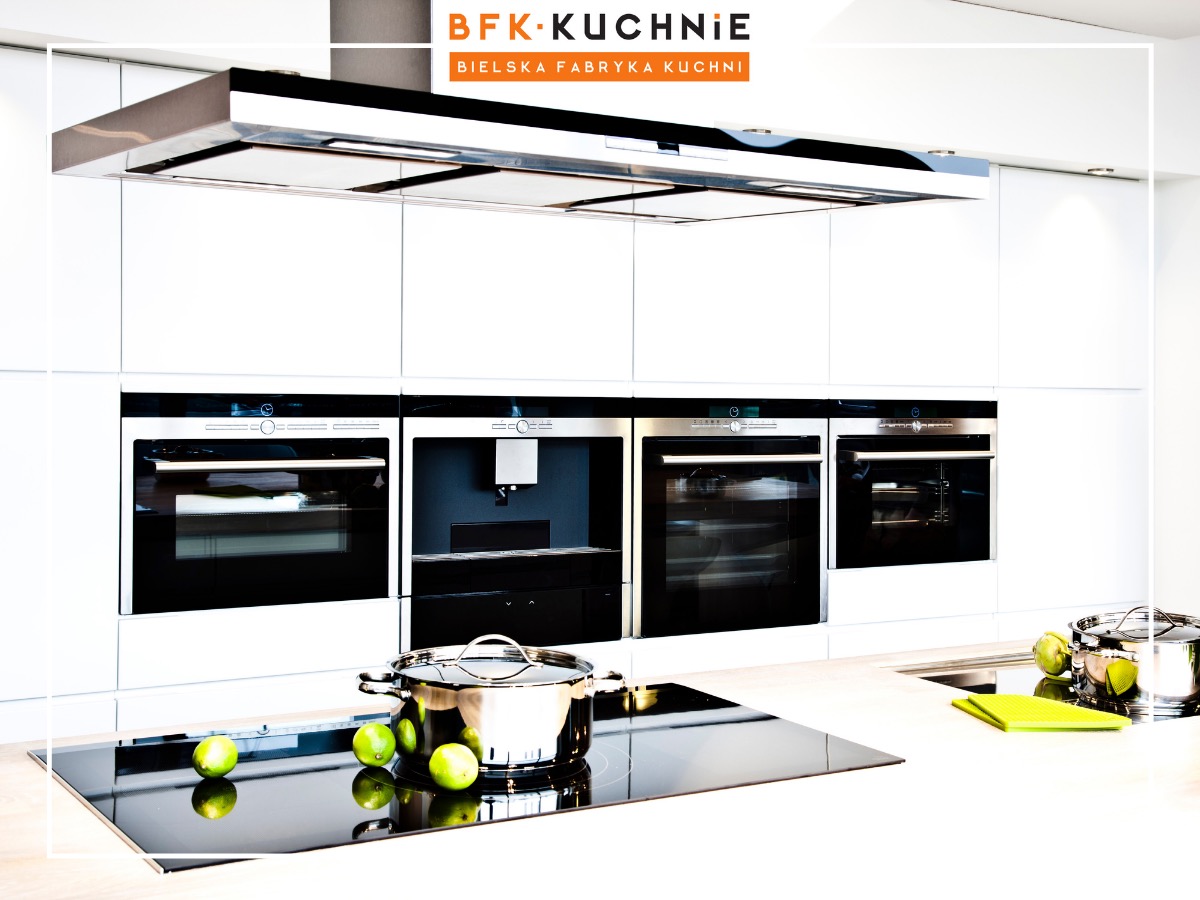 Minimalistyczna, biała kuchnia z zabudowanymi piekarnikami, ekspresem do kawy i płytą indukcyjną, na której leżą limonki i stoi garnek ze stali nierdzewnej.