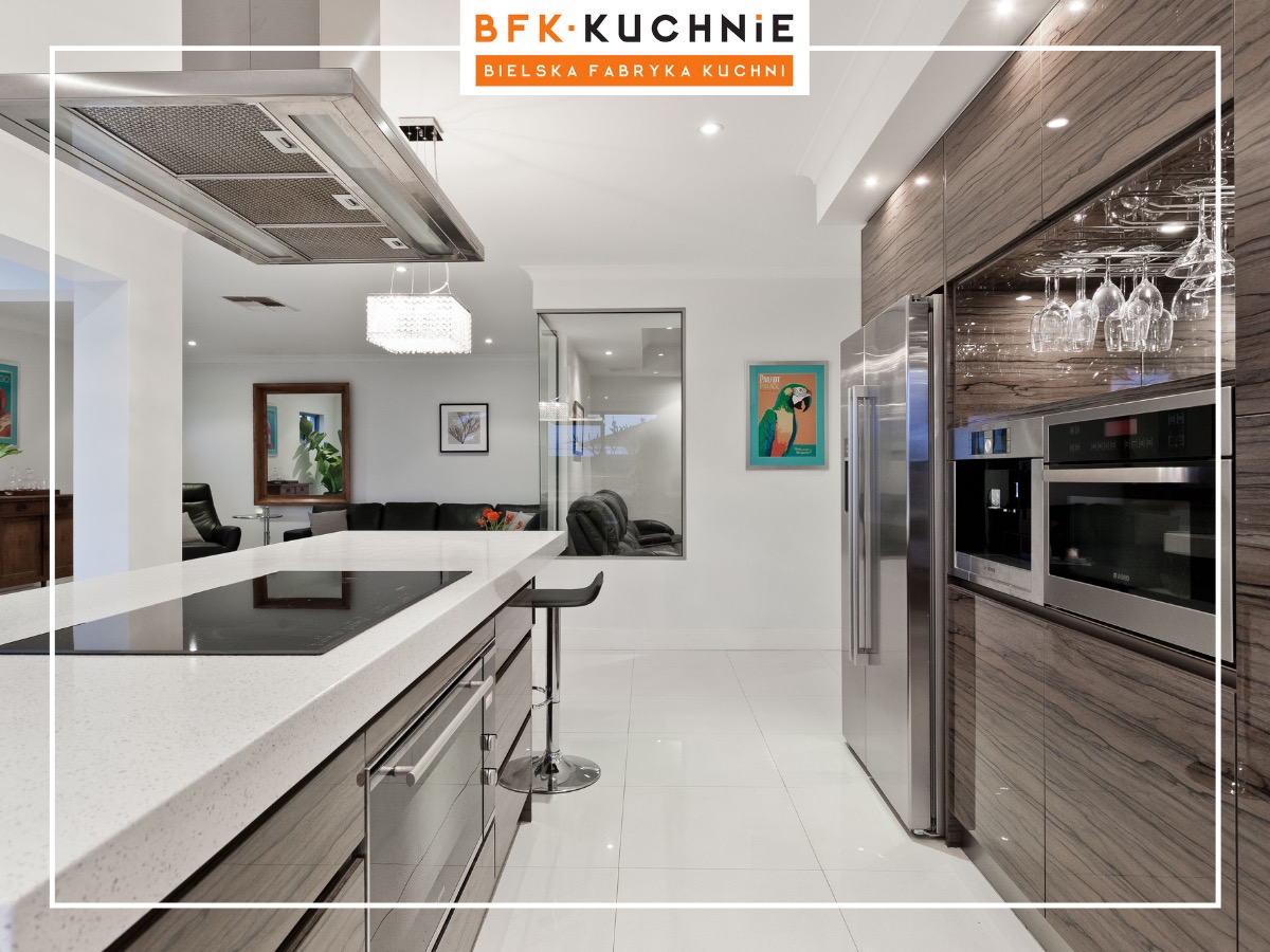 Nowoczesna kuchnia z wyspą i blatem z jasnego kamienia, fronty szafek w odcieniach szarości z wyraźnym rysunkiem drewna, zabudowa z wbudowanym sprzętem AGD, widoczne kieliszki zawieszone nad blatem...