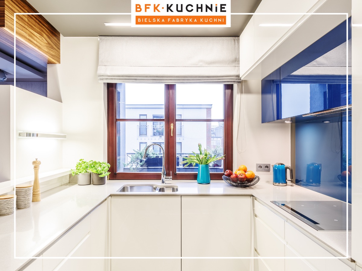 Jasna kuchnia w stylu minimalistycznym z drewnianą ramą okienną, widok na budynek z oknami, na blacie owoce w koszyku, niebieski czajnik i wazon z tulipanami.